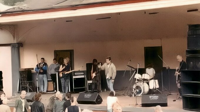 A photo of a punk band playing at an outside bandstand, a smattering of punks stand in the audience.
