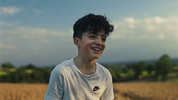 Lawland, a teenage boy with dark messy hair, stands in front of a pastoral scene of wheat, trees, and blue clouded skies