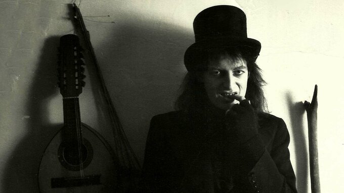 A dark black and white photo of a man sat next to a guitar. He's dressed in a goth style with a top-hat and long hair.