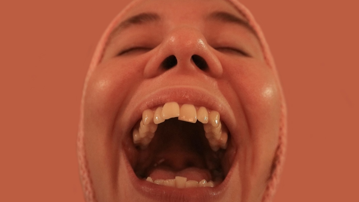 A close up of a person's face, their mouth is wide open and their eyes closed. The background and lighting is orange.