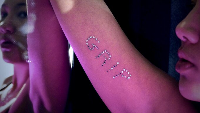 A close up photo of a woman's arm with 'Grip' bedazzled on it. Lit by pink and purple light.