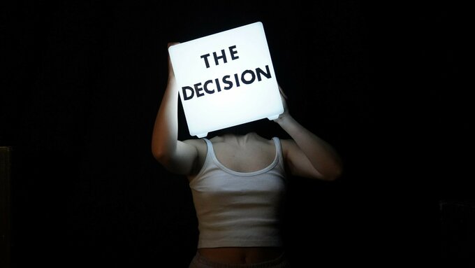 A headless woman wearing a shirt and pin skirt. Over her head is a light cube with the words ‘the decision’ on it.
