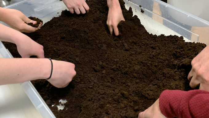 Several people's bare hands are digging in a box of soil in an indoor location.