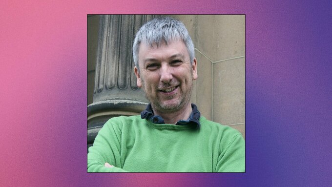 A headshot of Tom Murray,  smiles while standing behind a sandstone building. He is wears a green jumper and blue shirt.