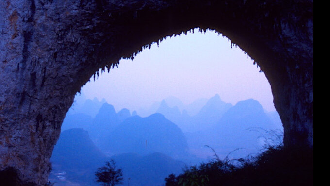Through a natural arch in the side of a mountain we see many layers of blueish hills and mountains, a Chinese landscape.