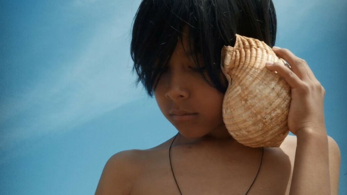 A child listens very carefully to a seashell. In the background there is a blue sky.
