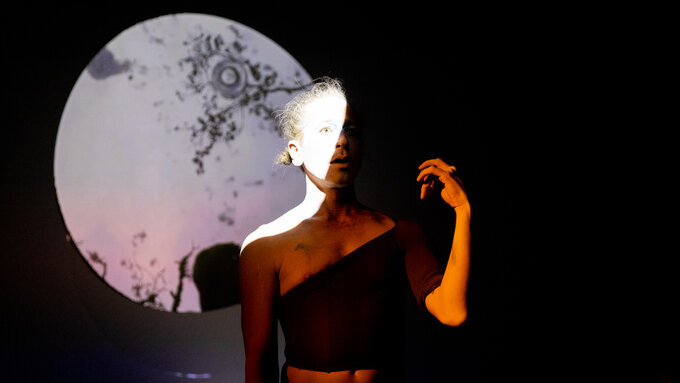 A dancer stands half in shadow, with one arm raised and fingers contracted. A full moon hovers in the background.