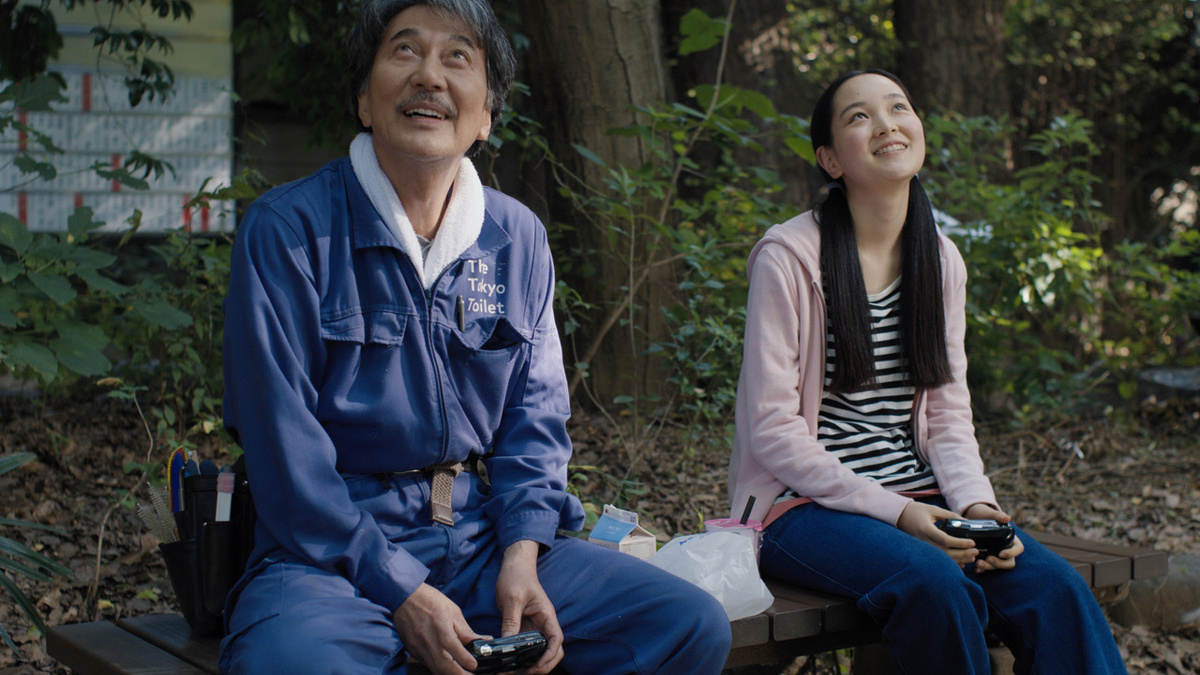 An older Japanese man sat with a younger Japanese woman on a bench in a wooded area. They both hold disposable cameras.