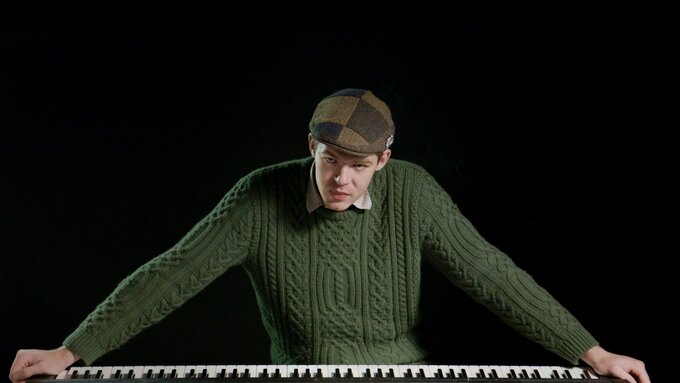 A man leaning over a keyboard, his arms spread wide across it. Wearing a green cord jumper and chequered brown flat cap.