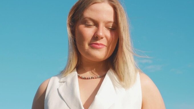 A photo of Kirsteen Harvey, a blonde white woman. She wears a white dress and is against a blue sky.