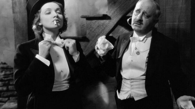 Marlene Dietrich wears a tuxedo and adjusts her bowtie whilst a man looks on.