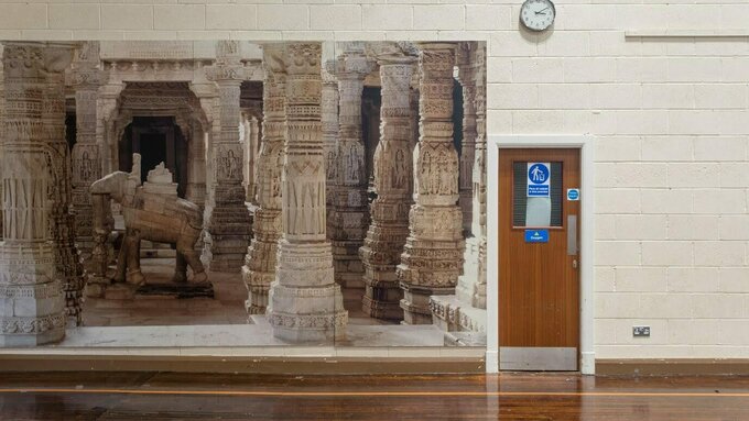 A wall of a gymnasium featuring a door, a clock and a large printed image of an ancient pillared building.