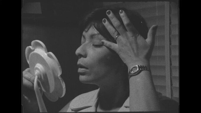 A black & white still of a woman touching up her eye make-up in a compact mirror.