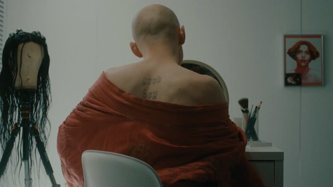 Someone sits at a dressing room table, with a draped blanket over their shoulders. A wig on a stand sits in the corner.