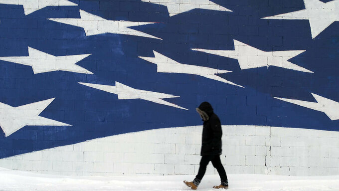 A hooded figure walks next to a wall with a massive mural of white stars on a blue background, like the USA flag.