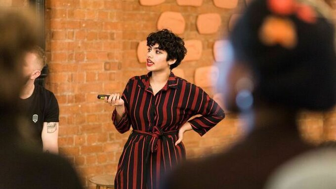 A photo of an Indian woman with curly short black hair, and red lipstick. Mid-speech, with a hand on her hip.