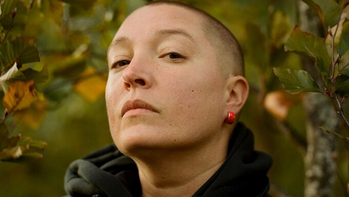 A white person with a shaved head with a red hoop earring and a black jacket and hoodie. There are leaves behind them.