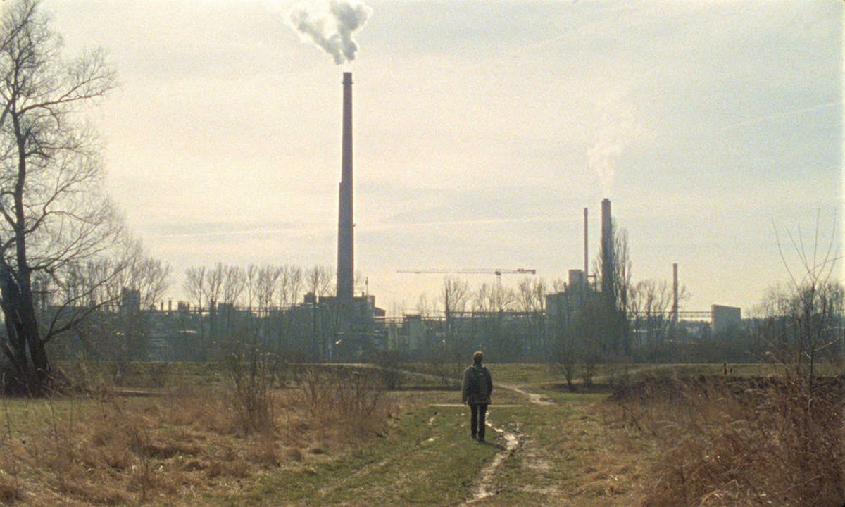 A young person walks through a forest towards the city on a sunny day. In the background are factory smokestacks.