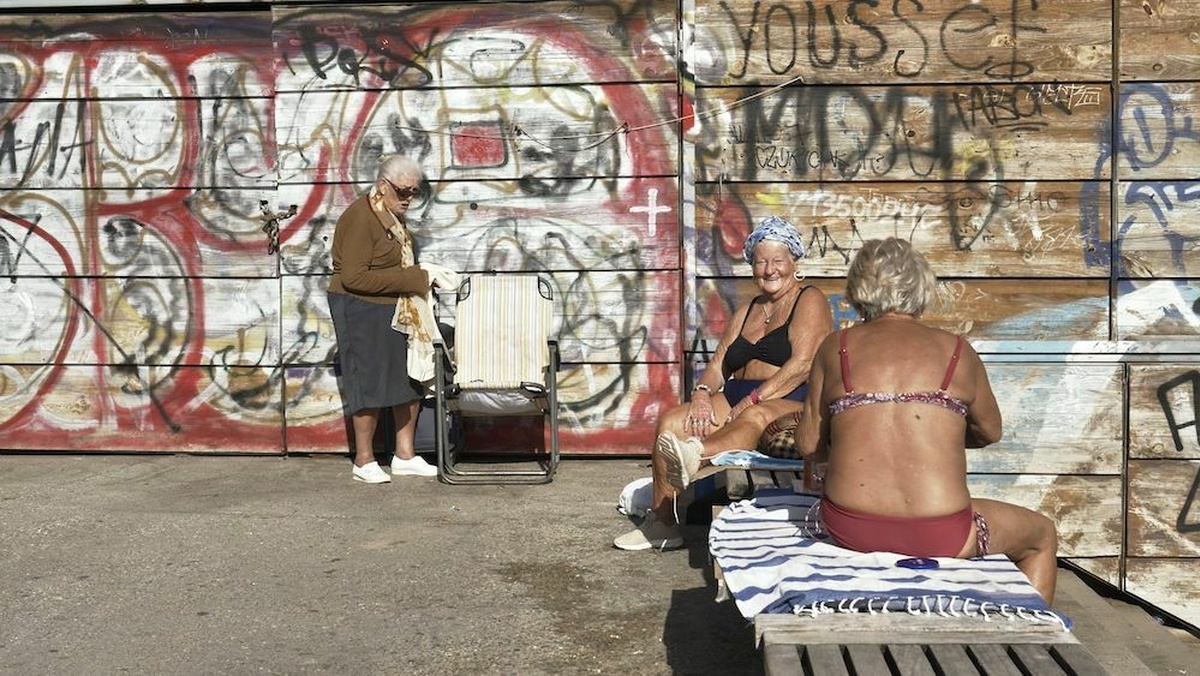 Three older women sunbathe. In the background you can see graffiti.