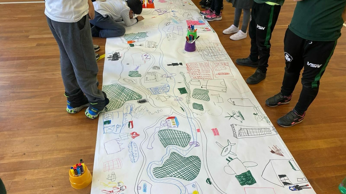 Several children stand around a large white scroll of paper with various drawings on it.