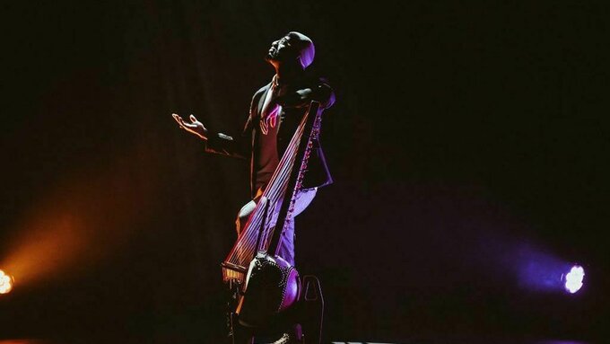 A man stood with his hands up to the sky holding a kora. A West African instrument reminiscent of a cello.