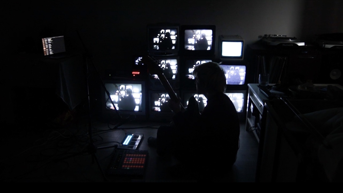 A person sitting cross-legged in a dark room. Behind them, a wall of flickering CRTs.