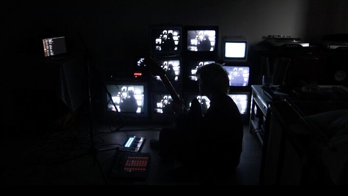 A person sitting cross-legged in a dark room. Behind them, a wall of flickering CRTs.