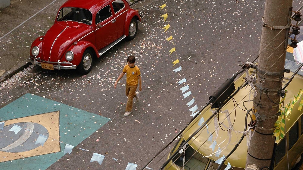 A boy wearing a Brazil shirt walks down an empty street. There is a painted Brazilian flag & floating football confetti.