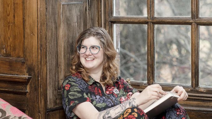 A woman sitting by a window with a notebook or book. She has glasses and a tattoo on her arm.