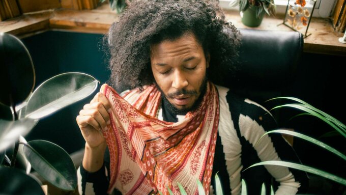 A slim black man sat on a sofa with his eyes closed serenely, surrounded by green plants.