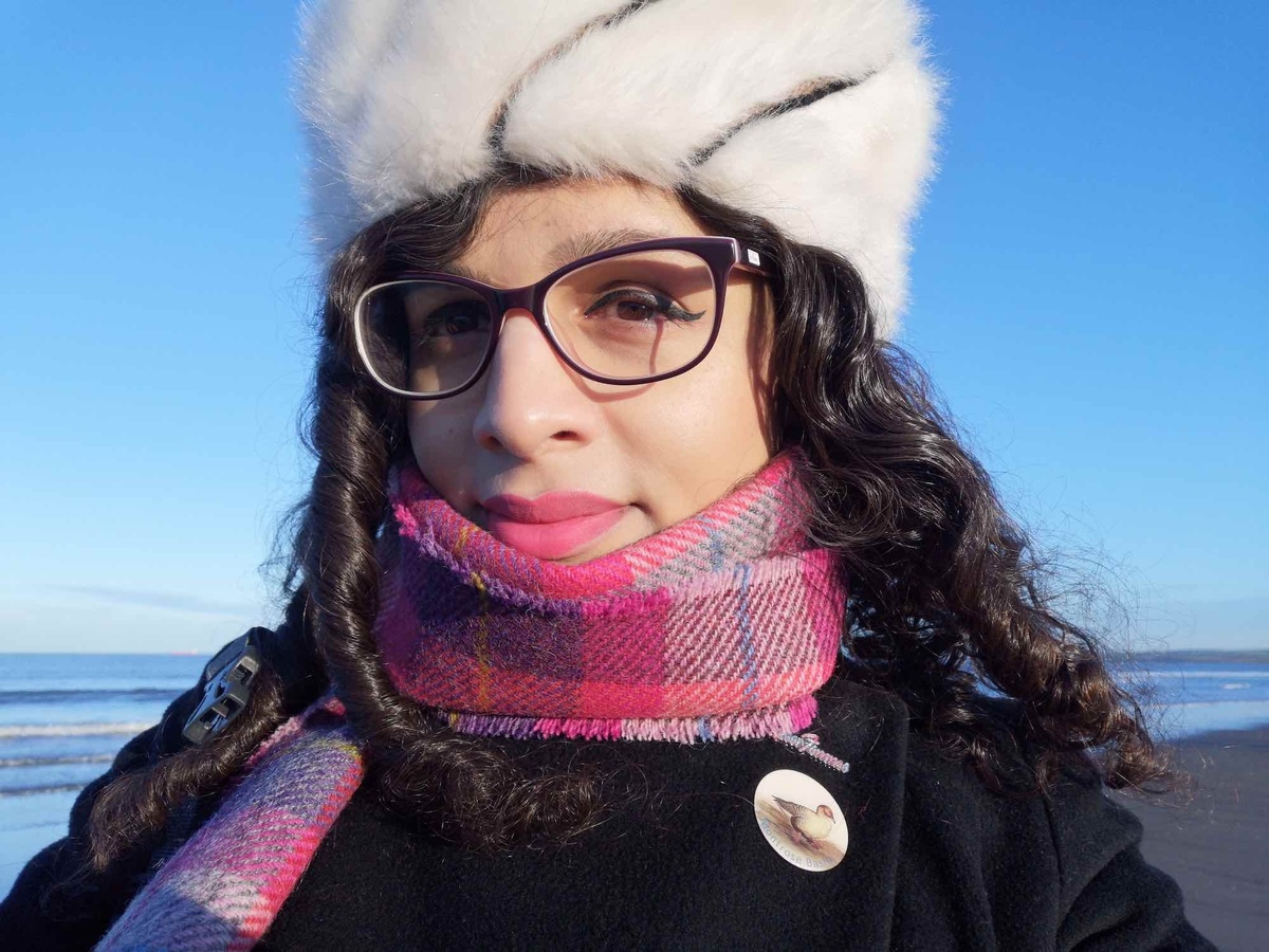 A photo of Nat Raha, a brown skinned woman wearing glasses, a thick hat, and scarf in front of a beach.