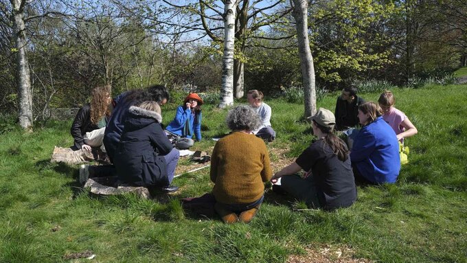 A group of people sitting in a circle in a sunny outdoor forested setting.