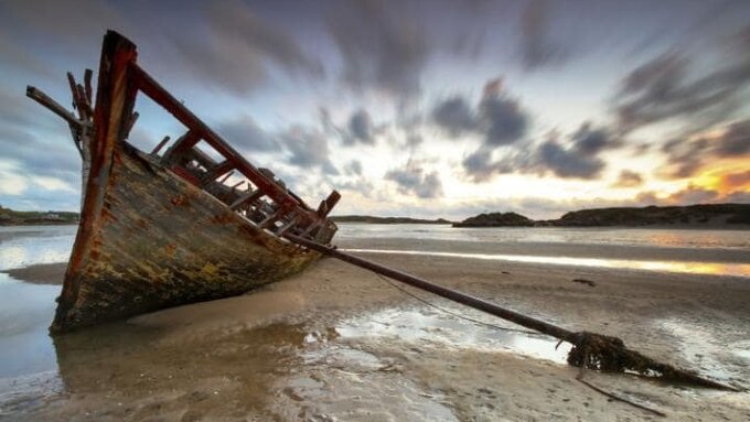 A view of a beach at sunset with a decayed wooden ship resting on the sands.