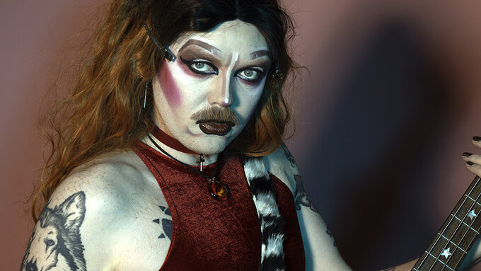 A person in heavy drag makeup with long highlighted hair and a moustache.