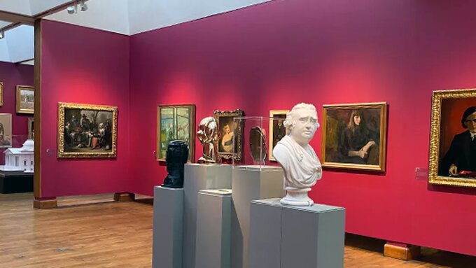 A museum gallery space, a white marble bust of an older man sits in front of gold framed paintings hung on red walls.
