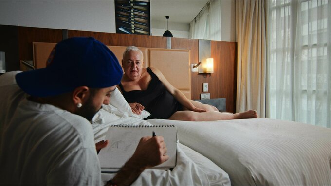 A person lays across a bed in a hotel room. They are looking at someone who is sketching them.