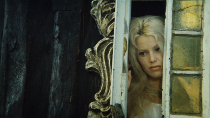 A white woman with bleach blonde hair and dark roots, peeking through a faded baroque gold framed window.
