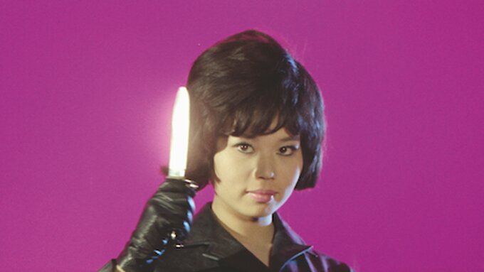 Against a pink background a woman holds up a knife to the camera.