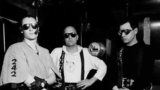 A photo of three men in out-there club attire including quilted chaps, climbing gear and dark sunglasses.