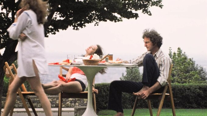 Two women and a man sit outside and eat food, one woman has got up to leave while the man writes