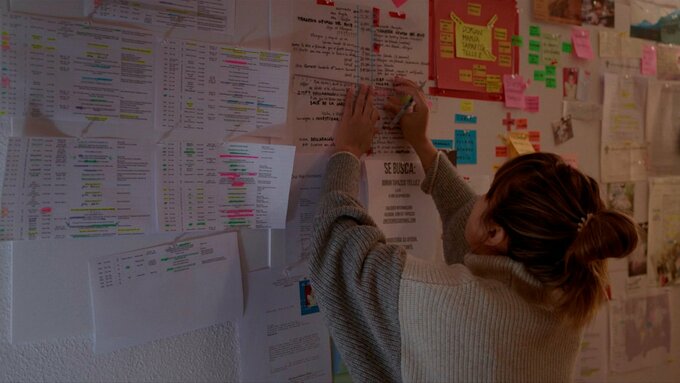 A woman is writing notes on a piece of paper stuck to a wall which is covered in other pieces of paper.
