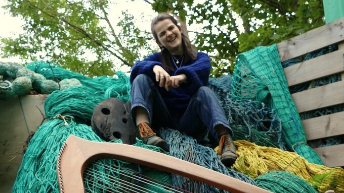 Grace is sitting on a pile of bright green fishing nets with a dark blue knitted jumper on and blue jeans.