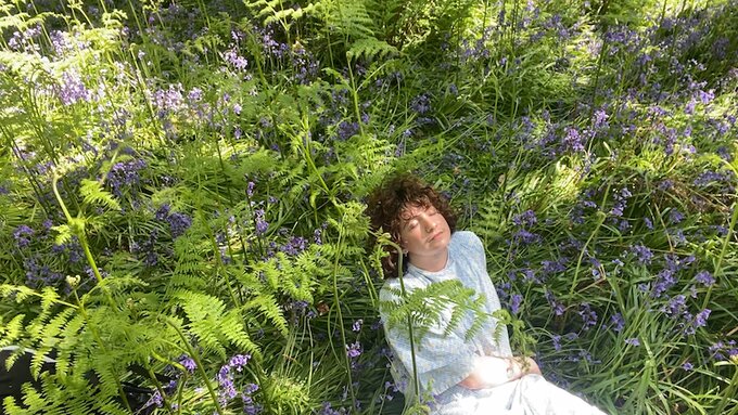 A man with long, curly brown hair sits leaning back in a field of bluebells. He wears a hospital gown.