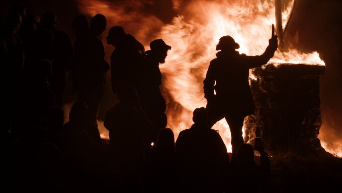 A dramatic nighttime scene features silhouetted figures gathered around a large, intense fire.