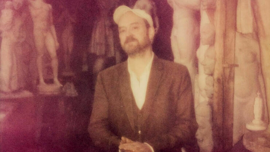 A grainy photo of a bearded man wearing a suit and baseball cap stood surrounded by neoclassical sculptures.