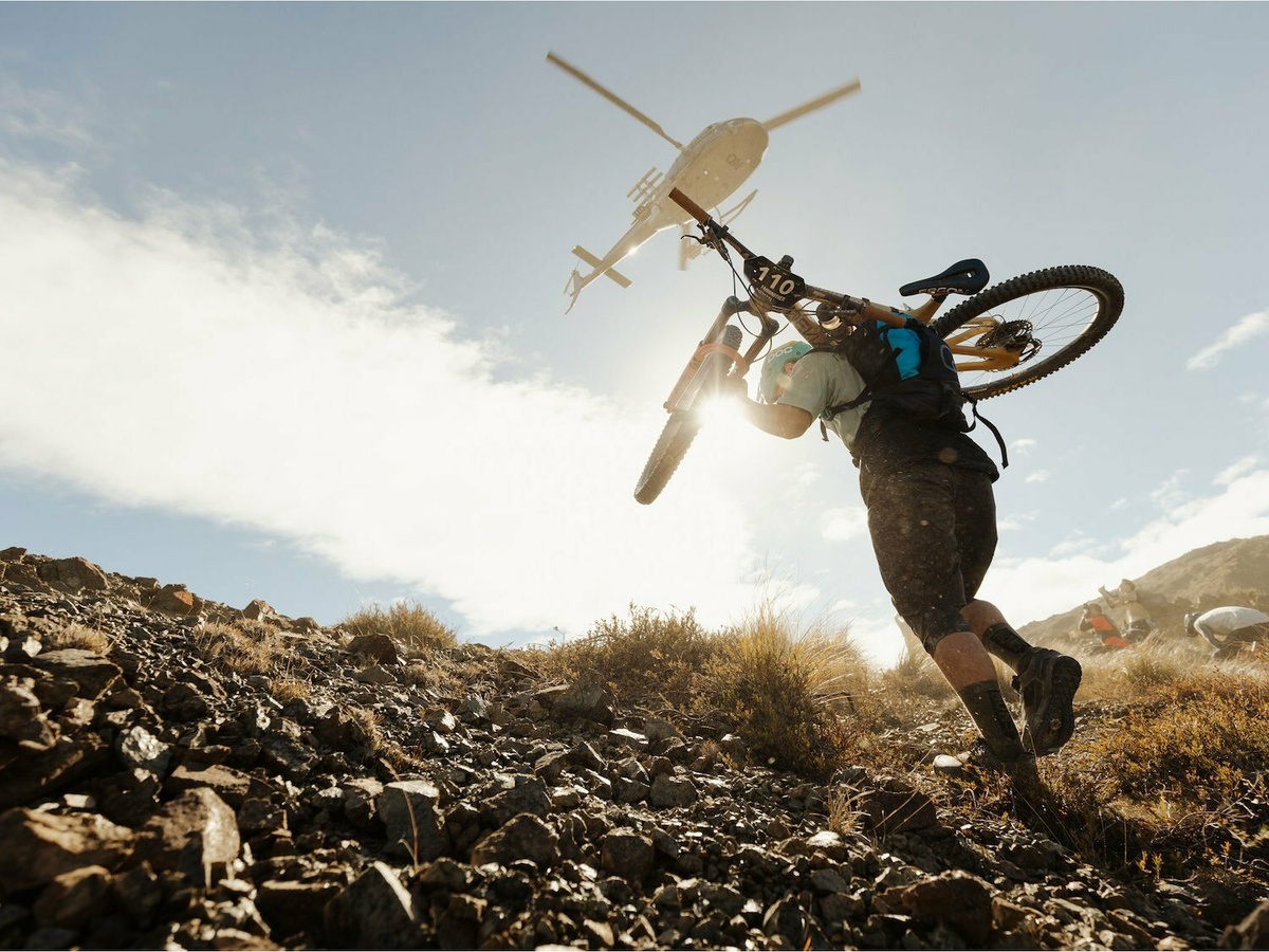 A person carrying a bike up a rocky hill, a helicopter flies overhead.