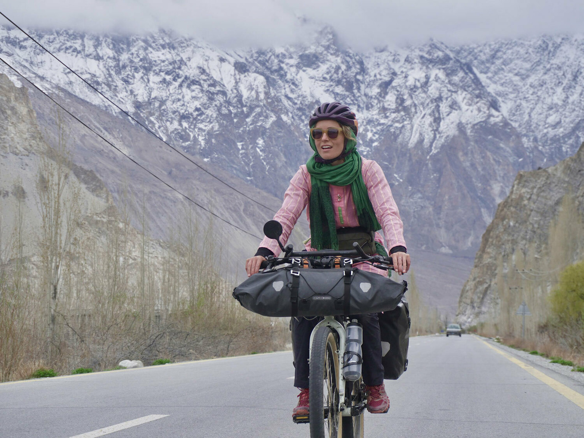 A woman cycling down a road, snow peaked mountains are visible in the distance.