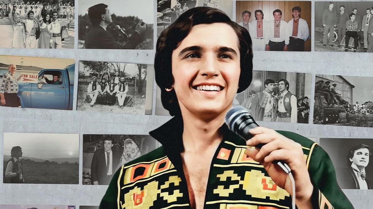 A smiling man with dark brown hair and light skin holds a mic. He wears a loud black and gold patterned shirt.