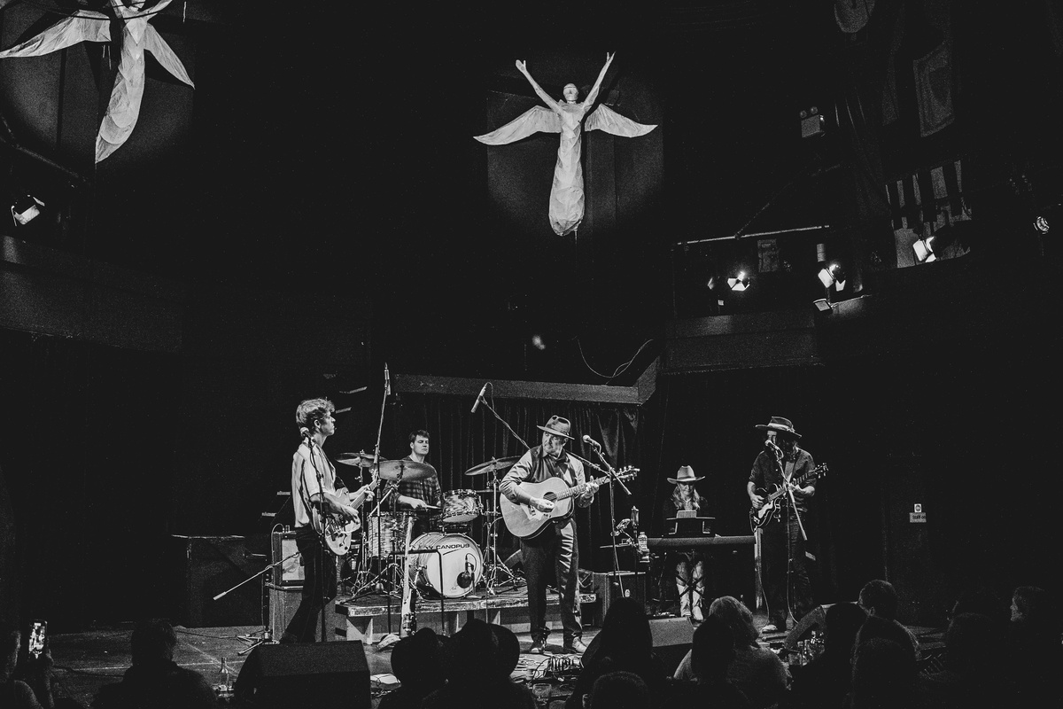 A black and white photo of Alan Fletcher and his band, two sculptures of angels appear to float above them.