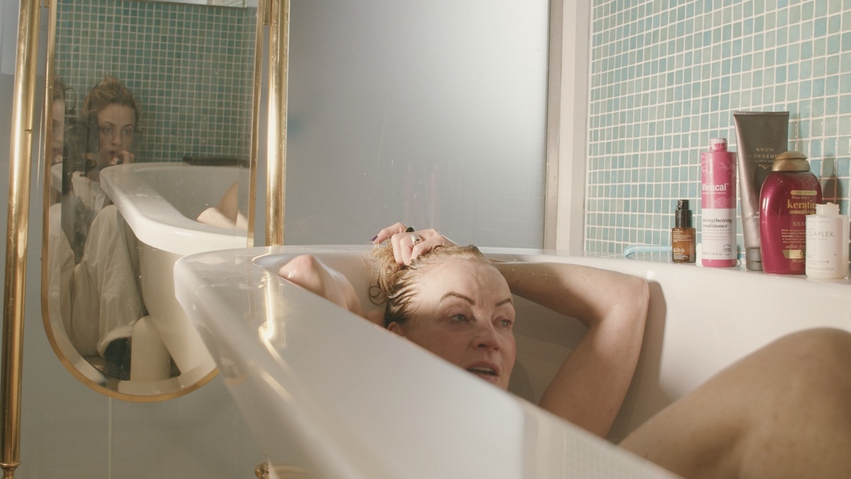 A woman sat in a bathtub. Another woman is reflected in a mirror, appearing to film them both.